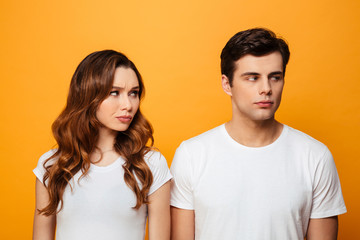 Photo of displeased boyfriend and girlfriend in white t-shirts being in fight while posing on camera with offended look, isolated over yellow background