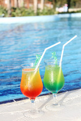Two glasses with colored cocktails are around the pool