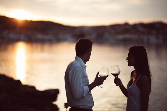 Two young people at the beach,making a toast for special occasion.Looking at the sun,enjoying the view with two glasses of wine.Drinking red wine.Celebration of anniversary.Birthday surprise.Honeymoon