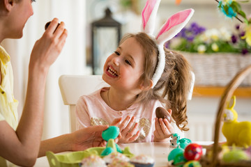 Mother and daughter celebrating Easter, eating chocolate eggs. Happy family holiday. Cute little girl in bunny ears laughing, smiling and having fun.