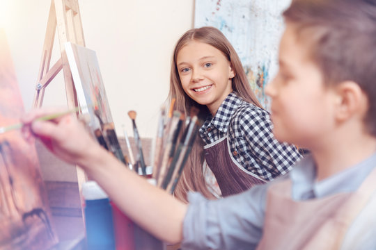 Cheerful atmosphere only. Young girl wearing an apron grinning widely sitting at an easel and posing for the camera.