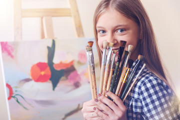 SEt of a real artist. Positive girl sitting at her easel and holding many already used painting brushes while smiling and posing for the camera.