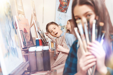 Wild and fun students. Male youngster joking while holding a painting brush while sitting next to his classmates and learning art.