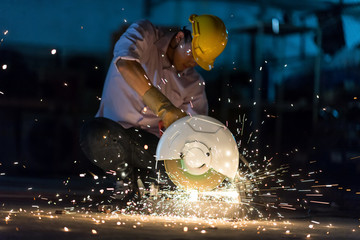 Low light image of Worker ware yellow helmet kneel use electric steel cutter machine. He has a Hard work in Factory or garage. Industry in construction site concept.