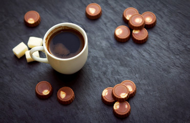 Cup of freshly brewed coffee, milk chocolate candies and sugar on black stone tray. Sweet breakfast or snack.