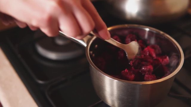Womans hand are holding bowl on a burner of cooker and mixing red berries. Preparation of cherry jam in home kitchen by housewife