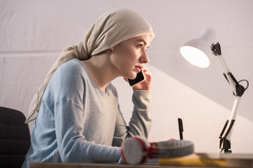 side view of young woman in kerchief talking by smartphone and taking notes