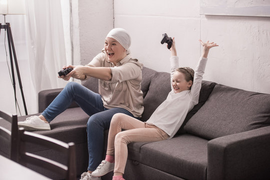 cheerful grandmother and granddaughter playing with joysticks, cancer concept