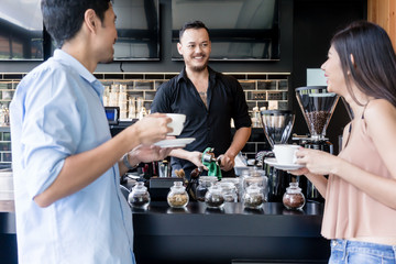 Friendly Asian young bartender cleaning the coffee maker while talking with two young customers over the bar counter of a modern cafeteria