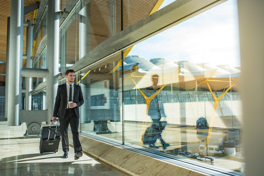 young businessman walking at the airport terminal with luggage smiling with a coffee