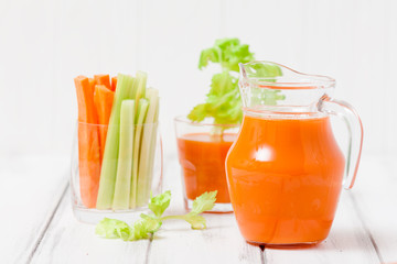Carrot juice in beautiful glasses, cut carrot and green celery on wood bark bowl on white wooden background. Fresh vegetable drink. Close up photography. Selective focus. Horizontal banner