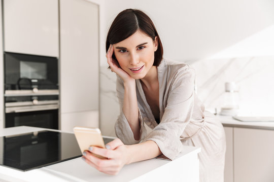 Photo of attractive woman with short dark hair wearing beautiful robe looking on camera with smile, while using smartphone in kitchen