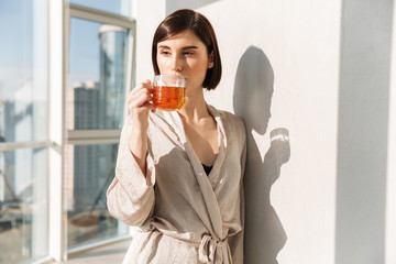 Gorgeous woman with short dark hair in robe drinking tea and enjoying sunny weather, while standing...