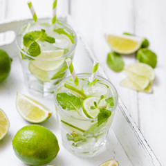 Caipirinha, mojito cocktail with lime, brown sugar, ice and mint leaves in beautiful glasses, cut green citrus on white wooden background. Summer alcohol drink. Close up photography. Selective focus