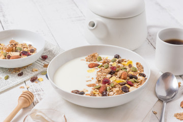 Cereal breakfast with dried almond, cranberry, grapes, Pistachios nuts and cornflakes over plain yoghurt in white bowl with honey, black coffee cup and spoon on white wooden background, healthy meal