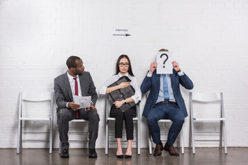 businessman covering face with card with question mark while waiting for job interview with multiethnic business people