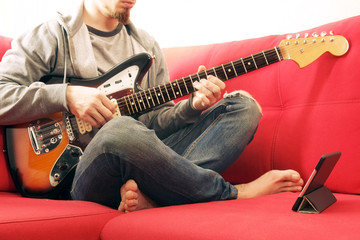 Obraz premium Casually dressed young man with guitar playing songs in the room at home. Online guitar lessons concept. Male guitarist practicing chord grips.