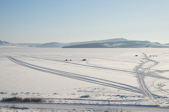 Winter lake with fishermen and traces of cars in the snow, against the background of mountains.