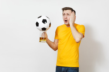 Sad upset young bewildered European man, fan or player in yellow uniform hold pint mug of beer, soccer ball cheer favorite football team isolated on white background. Sport, play, lifestyle concept.