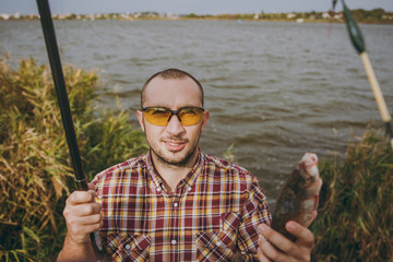 Young unshaven smiling man in checkered shirt and sunglasses pulled out fishing pole and holds...
