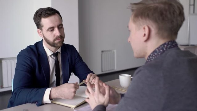 Man with beard listens to client in office at table to job in business. Serious businessman makes an interview with a male person and holds a pen over his notebook.
