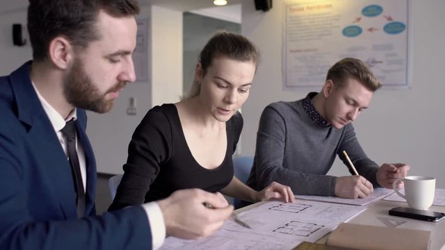 Three architects are discussing building blueprints in their office. Man blue suie, woman in dress and male enjineer in grey jumper are sitting in a row at the table with paper schemes and coffee cup