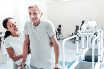 I am determined. Inspired old grey-haired man exercising while a pretty smiling dark-haired dark-eyed afro-american woman helping and holding him