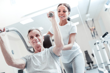 Helping her patient. Inspired old grey-haired man exercising on a training device and an attractive young dark-haired female trainer standing behind him and touching him