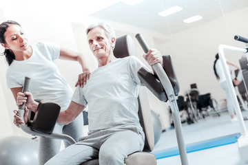 Fototapeta na wymiar Being vigorous. Exuberant old crippled grey-haired man smiling and exercising on a training device and a beautiful young female trainer standing near him and touching him on the shoulder