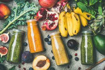 Flat-lay of colorful smoothies in bottles with fresh tropical fruit and vegetables on concrete background, top view. Healthy, clean eating, vegan, vegetarian, detox, dieting breakfast food concept