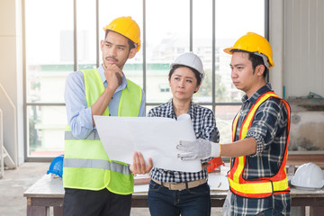 Fototapeta na wymiar Serious Three Asian male and female architect engineers with helmeted thinking about they project and holding plan in paper standing at construction site and glass frame background.