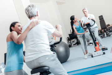 Perfect. Smiling old grey-haired man exercising on a training device and looking in the mirror while a happy young dark-haired afro-american female trainer smiling and touching him