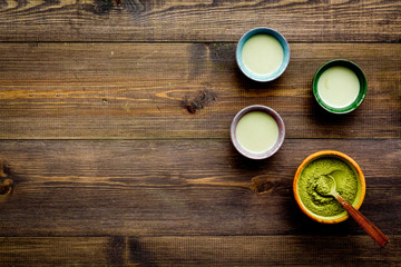 Brew matcha tea. Bowl with powder and cups with beverage on dark wooden background top view copy space