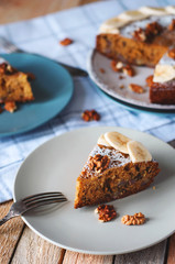 Angle view of sliced banana cake with powdered sugar and walnut on plate with fork. Cozy still life in light tones on wooden background with checked kitchen towel.