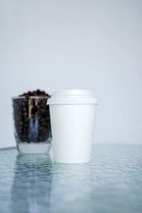 A white paper glass (one-time) for coffee and coffee beans in transparent glass is on the table. Background is blurred, white and blue. Place for the title on the glass. Side view with copy space