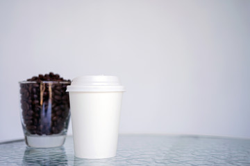 A white paper glass (one-time) for coffee and coffee beans in transparent glass is on the table. Background is blurred, white and blue. Place for the title on the glass. Side view with copy space