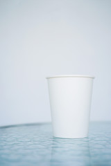 A white paper glass (one-time) for coffee is on the table. Background is blurred, white and blue. Place for the title on the glass. Side view with copy space