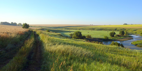 Sunny summer landscape with ground country road.Green hills,fields and meadows.River Upa in Tula region,Russia.Sunrise.Quiet morning.Calm.Warm sunlight.Cloudless clear blue sky. 