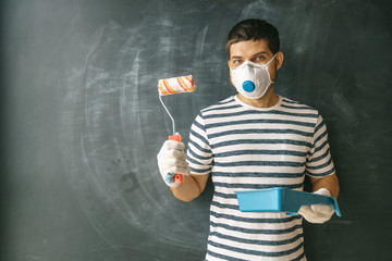 Portrait of a cheerful young master wearing a protective mask looking at the camera with a roller and paint in his hand on a dark background