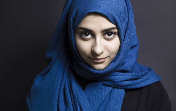 Portrait of a young Muslim woman wearing a blue hijab looking directly at the camera on a dark background. Diversity and modern Islamic culture concept.