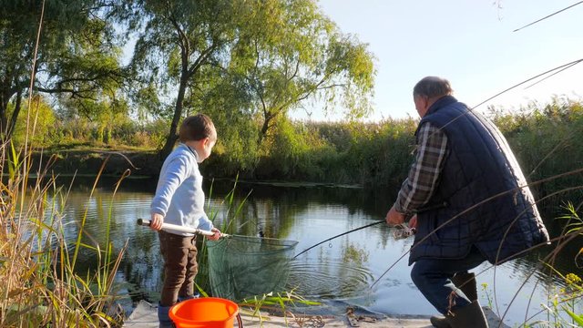 happy childhood, fishing of grandson with grandfather on lake in spring on weekends among trees and grass