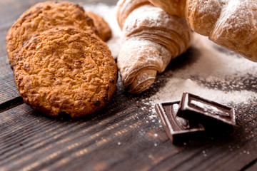 croissants sprinkled with powdered sugar and oatmeal cookies on a dark wooden table