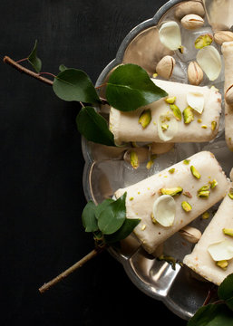 Pista kulfi, Indian ice cream on serving plate with ice cubes, pistachios, rose petals, tree branches with green leaves on black background, top view. Traditional Rajasthani Indian cuisine.
