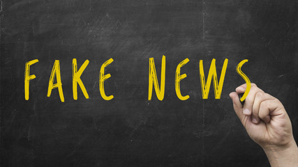 Male hand writing the words Fake News on chalkboard as a reminder to be aware of hoaxes and...