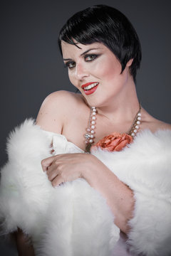 Brunette girl with short hair, French mane in the style of the 20s. She wears white fur coat and jewelry