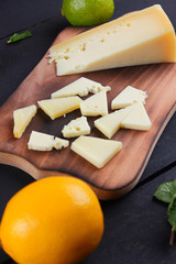 Hard yellow cheese, lemon, lime and fresh mint on a wooden black background, slices of cheese on a black background, top view, mint leaves with cheese with holes, rustic style, citrus, healthy food, v
