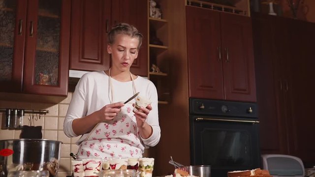 Young cook woman is creating authors desserts in a home kitchen. She is taking cup and leveling cream