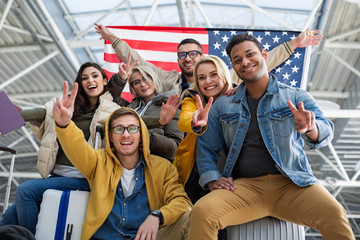 Low angle of happy men and women standing at the airport with banner of the united states and showing two finger gesture © Yakobchuk Olena