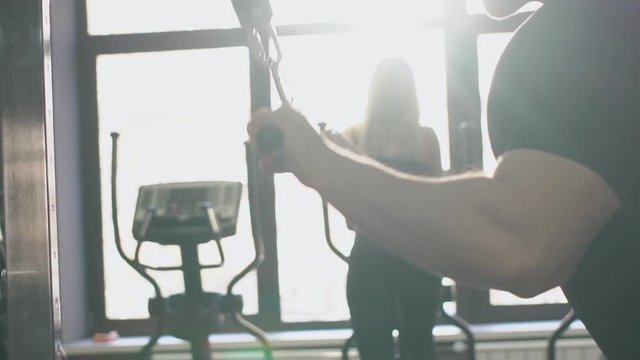 Arms of sportsman, who is doing triceps pushdown in sunny gym, close up. Muscular hands of male athlete are holding the cable equipment and silhouette of sportswoman, who is doing exercises on
