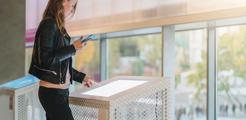 Young woman stands indoor and touches digital display while holding smartphone in her hand. Hipster...